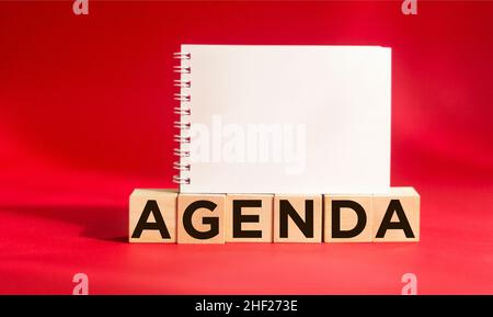 Agenda-Symbol. Holzblock mit dem Wort Agenda auf rotem Hintergrund mit einem Notizblock zum Schreiben. Geschäfts- und Agendakonzept. Speicherplatz kopieren. Stockfoto