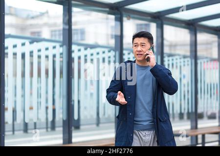 Ein Mann, der am Telefon sprach, ein fokussierter und ernsthafter Asiatisch an einer Haltestelle des öffentlichen Nahverkehrs, ein Geschäftsmann in der Nähe des Bürozentrums Stockfoto