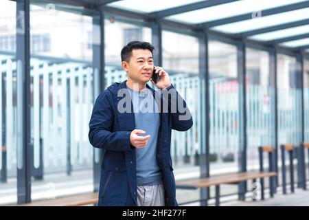 Ein Mann, der am Telefon sprach, ein fokussierter und ernsthafter Asiatisch an einer Haltestelle des öffentlichen Nahverkehrs, ein Geschäftsmann in der Nähe des Bürozentrums Stockfoto