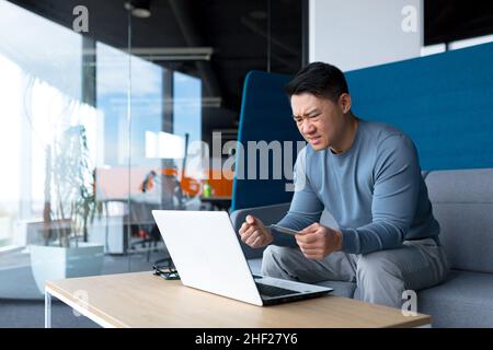 Verärgerte asiatische Mann versucht, Banking mit Laptop und Kreditkarte, Geschäftsmann in der Arbeit in modernen Büro zu tun Stockfoto