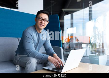 Porträt eines männlichen Lehrers, Asiaten, die vom Büro des Schulungszentrums aus arbeiten, mit einem Laptop aus der Ferne, mit Blick auf die Kamera und lächelnd Stockfoto