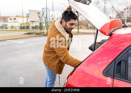 Junger Mann, der den Rucksack in den Kofferraum des roten Autos auf den Parkschlitz nimmt oder steckt Stockfoto