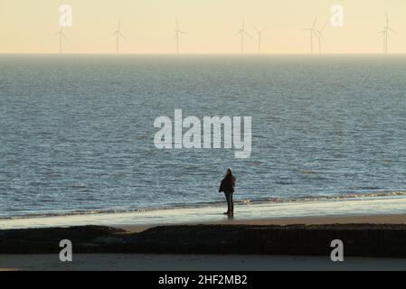 Mann, der alleine am Strand steht und auf dem Meer in Frinton in Essex mit dem Windpark Gunfleet Sands am Horizont blickt Stockfoto