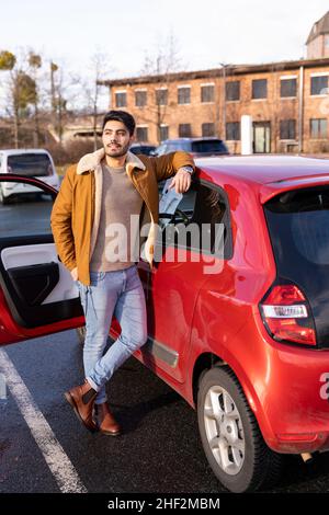 Ein lateinischer oder arabischer Mann lehnte sich neben dem Auto mit geöffneter Tür auf dem Parkplatzplatz der Stadt an Stockfoto