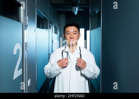 Porträt eines ernsthaften asiatischen Arztes, eines Mannes in der Klinik, im Korridor in der Nähe der Stationen, der aufmerksam auf die Kamera starrte Stockfoto