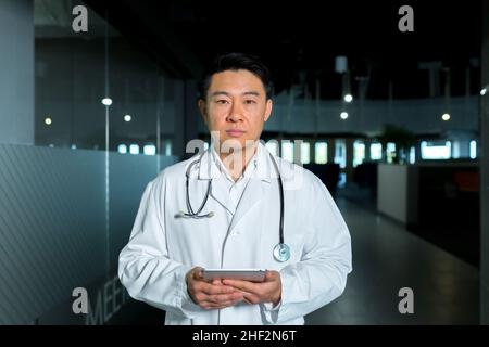 Konzentriert und selbstbewusst blickt der asiatische Arzt im Gang der Klinik auf die Kamera und hält einen Tablet-Computer in der Kamera Stockfoto