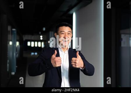 Erfolgreicher Geschäftsmann freut sich, wenn er die Kamera anschaut, zeigt Daumen nach oben und Schilder in Ordnung, asiatischer Boss glücklich und lacht Stockfoto