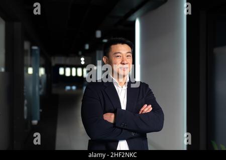 Porträt eines erfolgreichen asiatischen Manager-Chefs, mit gekreuzten Armen in einem Business-Anzug, der pensiver auf die Kamera schaut, ernsthafter Mann im Büro Stockfoto
