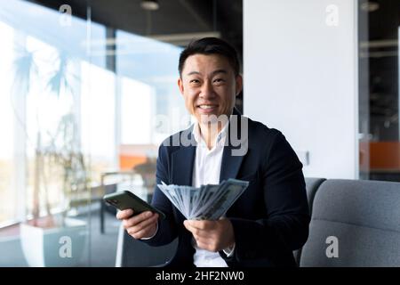 Erfolgreicher asiatischer Chef-Geschäftsinhaber, schaut auf die Kamera und freut sich, feiert den Sieg und die erhaltene Leistung, hält der Mann ein Telefon und cas Stockfoto