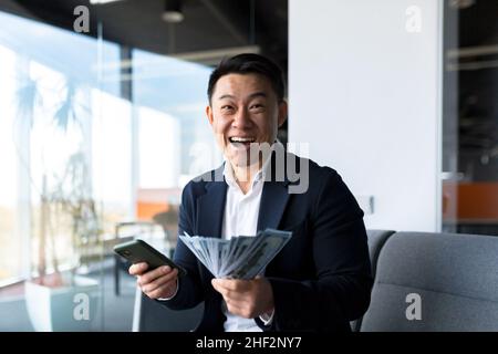 Erfolgreicher asiatischer Chef-Geschäftsinhaber, schaut auf die Kamera und freut sich, feiert den Sieg und die erhaltene Leistung, hält der Mann ein Telefon und cas Stockfoto