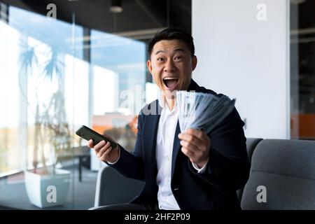 Erfolgreicher asiatischer Chef-Geschäftsinhaber, schaut auf die Kamera und freut sich, feiert den Sieg und die erhaltene Leistung, hält der Mann ein Telefon und cas Stockfoto