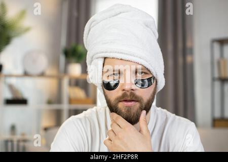 Nahaufnahme eines gutaussehenden, fröhlich lächelnden kaukasischen Mannes mit weißem Handtuch auf dem Kopf und schwarzen Flecken unter den Augen, der die Hand auf sein Kinn legte und die Kamera ansah Stockfoto
