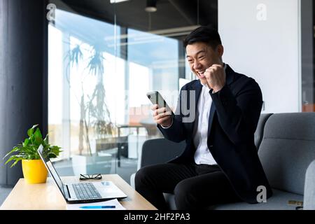 Erfolgreicher asiatischer Geschäftsmann feiert den Sieg, schaut glücklich ins Telefon und freut sich im modernen Büro zu sitzen Stockfoto