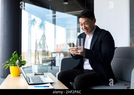 Erfolgreicher asiatischer Geschäftsmann feiert den Sieg, schaut glücklich ins Telefon und freut sich im modernen Büro zu sitzen Stockfoto