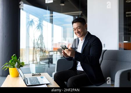 Erfolgreicher asiatischer Geschäftsmann feiert den Sieg, schaut glücklich ins Telefon und freut sich im modernen Büro zu sitzen Stockfoto