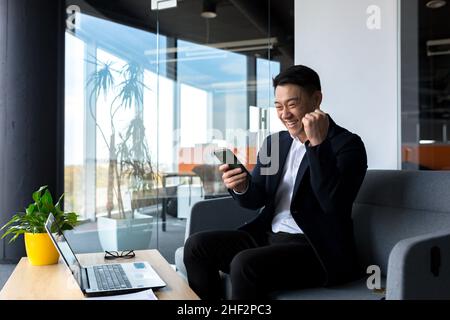 Erfolgreicher asiatischer Geschäftsmann feiert den Sieg, schaut glücklich ins Telefon und freut sich im modernen Büro zu sitzen Stockfoto