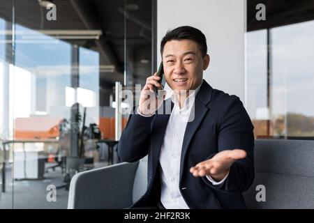 Asiatischer Geschäftsmann lächelt und freut sich glücklich am Telefon zu sprechen, freiberuflicher Mann im modernen Büro Blick auf die Kamera Stockfoto