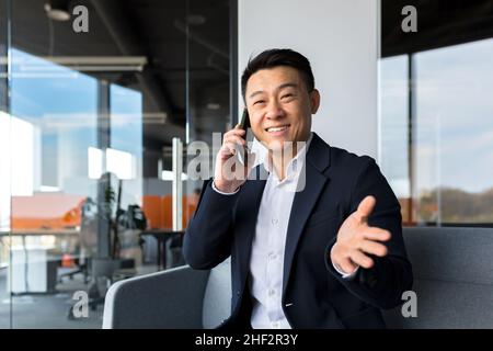 Der erfolgreiche chinesische asiatische Geschäftsmann blickt auf die Kamera und lächelt am Telefon Stockfoto