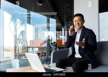 Asiatischer Geschäftsmann lächelt und freut sich glücklich am Telefon zu sprechen, freiberuflicher Mann im modernen Büro Blick auf die Kamera Stockfoto