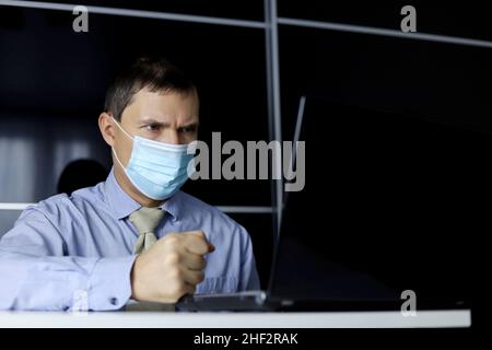 Wütender Mann mit Gesichtsmaske und Bürokleidung, der seine Faust auf den Tisch schlug, während er am Laptop saß. Konzept eines verärgerten Chefs während eines Online-Meetings Stockfoto
