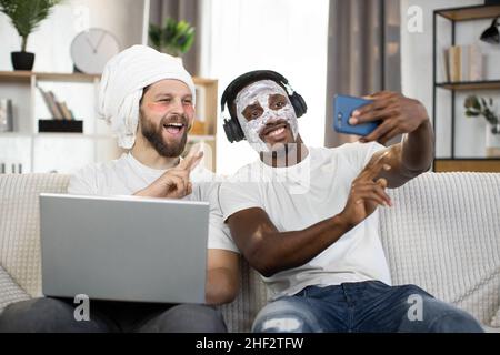 Männer Hautpflege zu Hause. Zwei junge, multiethnische Männer, die Gesichtshygieneverfahren durchführen, verwenden Augenaufnäher und Gesichtsmaske, um Selfie-Fotos auf dem Smartphone zu machen. Schwule Paare zu Hause Stockfoto