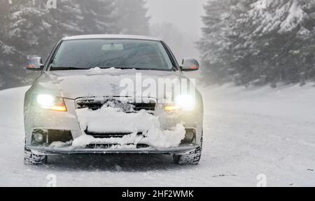 Silbernes Auto auf schneebedeckter Winterstraße geparkt, verwackelte Bäume Hintergrund, Vorderansicht, Scheinwerfer leuchten Stockfoto