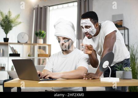 Attraktive fröhliche junge multirassische zwei männliche Freunde mit Augenflecken und Gesichtsmaske lachen, wenn sie lustige Videos oder Fotos am Computer zu Hause anschauen. Männer Hautpflege zu Hause Stockfoto