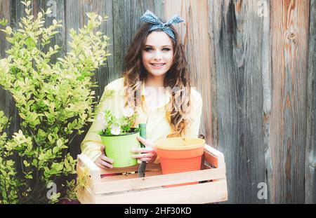 Ziemlich lächelndes Mädchen hält Gartengeräte auf hölzernen Hintergrund. Pflanz- und Blumenanbaukonzept. Die Gärtnerin kümmert sich um ihren Hinterhof Stockfoto
