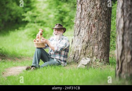 Glücklicher Mann, der einen frisch gepflückten Pilz in der Hand hält. Sammeln Von Wildpilzen. Großvater mit einem Korb von Pilzen und einem überraschenden Gesichtsausdruck. Stockfoto