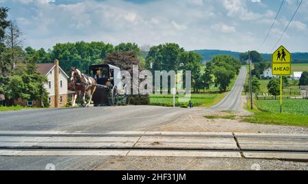 Amish Horse und Buggy nähern sich an einem sonnigen Tag auf einer Landstraße, die die Schienen überquert Stockfoto