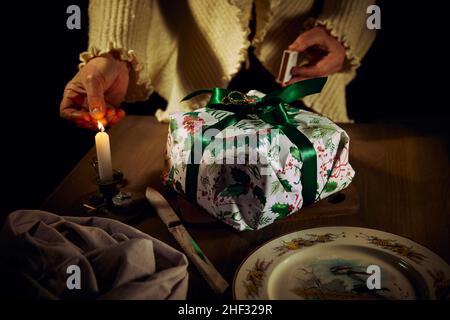 Eine Frau zündet eine Kerze auf einem Holztisch an und legt einen Weihnachtskuchen in eine Geschenkbox, ein Messer, eine Serviette und einen Teller. Stockfoto