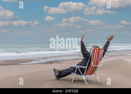 Erwachsener Mann, der mit den Armen in der Luft auf einer Strandliege vor dem Meer liegt Stockfoto