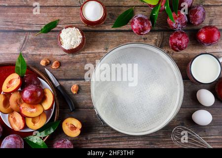 Backprozess Zutaten Eier, Schneebesen, Pflaumen und Backform auf rustikaler Holzoberfläche. Stockfoto