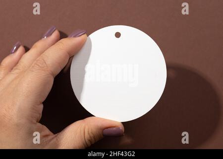 Frau Hand hält Pappe weißen weißen weißen weißen weißen Tag der runden Form mit wenig Loch für Kleidung im oberen Teil in der Mitte des braunen Hintergrund. Anhänger im Modell Stockfoto