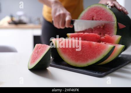 Eine Frau in der Küche schneidet eine Wassermelone, die Hände aus der Nähe, verschwommen. Saftige rote Fruchtbeere, vegetarisches Essen Stockfoto