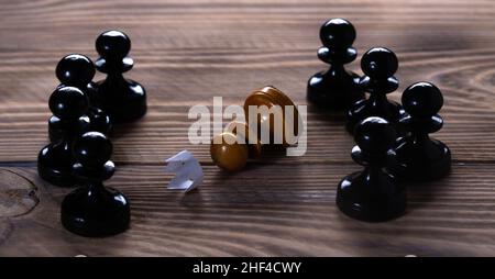 Eine einfarbige weiße Spielfigur mit einer abgelegten Papierkrone liegt umgeben von vielen schwarzen Spielfiguren. Besiegt. Stockfoto