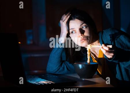 Mädchen isst Nudeln und arbeitet auf einem Laptop. Stockfoto