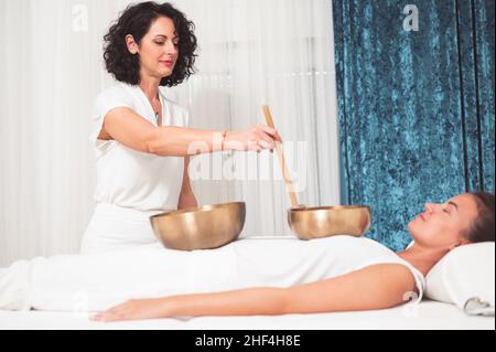 Schöne weibliche Therapeutin, die eine Klangheiltherapie über einer hübschen Frau praktiziert, die auf dem Tisch mit Klangschalen auf ihrem Körper liegt Stockfoto