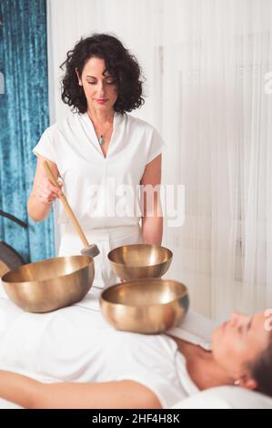 Schöne weibliche Therapeutin, die eine Klangheiltherapie über einer hübschen Frau praktiziert, die auf dem Tisch mit Klangschalen auf ihrem Körper liegt Stockfoto