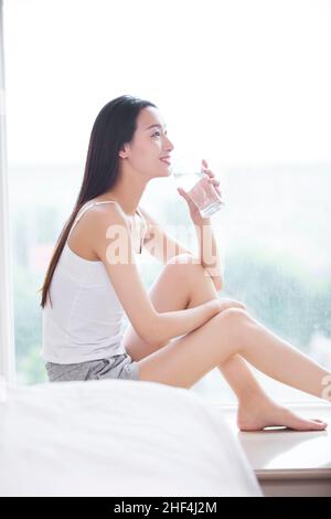 Junge Frau, die auf der Fensterbank sitzt und Wasser trinkt Stockfoto