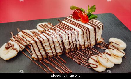 Nahaufnahme eines köstlichen Stückes hausgemachtem Käsekuchen mit Schokolade und Früchten auf Schiefer und rotem Hintergrund. Kochen Dessert und Kuchen Konzept Stockfoto