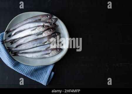 Frischer blauer Wittling auf Teller auf schwarzem Holzhintergrund. Frischer Fisch. Speicherplatz kopieren. Draufsicht. Stockfoto