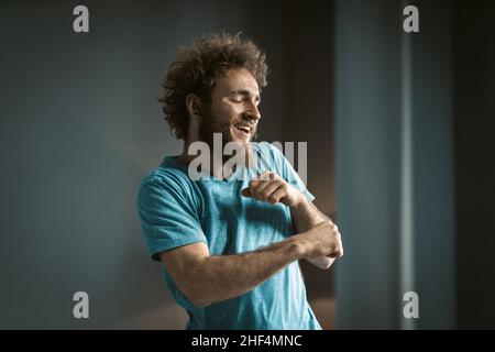 Coily Guy in einem blauen T-Shirt und einem Tattoo am Hals macht lustige Bewegungen an seinen Händen. Junger Mann tanzt im Zimmer zu Hause. Nahaufnahme. Hochwertige Fotos Stockfoto