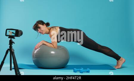 Pilates-Trainer erklärt Plank-Training auf Fitness-Toning-Ball, Aufnahme auf Kamera. Athletische Frau Dreharbeiten Trainingsstunde und mit Gymnastikgeräten, Unterricht Sportpraxis online. Stockfoto