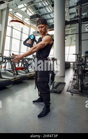 Ein junger Mann benutzt und genießt die Schlagmassagepistole, um sich zu entspannen und die Schmerzen im Körper und in den Muskeln im Fitnessstudio zu lindern. Stockfoto