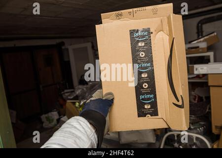 Frankfurt, Deutschland - 10. Dez 2021: Männliche Hand im Schutzhandschuh, der leicht beschädigt im Lager Amazon Cardboard Box hält Stockfoto