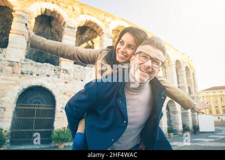 Glückliches Senior-Paar in der Liebe Spaß vor dem berühmten Denkmal von Italien. Blick aus der unteren Perspektive auf eine fröhliche Frau, die die Arme ausstreckt, während sie darauf sitzt Stockfoto