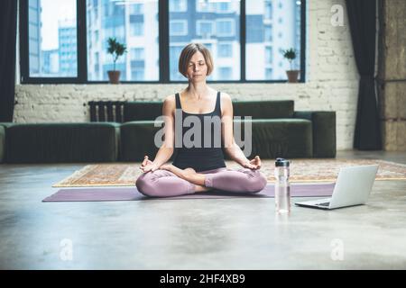 Die sportliche Frau sitzt auf einer Sportmatte und hat während der Quarantänezeit einen Online-Video-Chat mit ihrem Fitness Coach auf dem Laptop. Home Workouts. Stadthintergrund. Nahaufnahme. Hochwertige Fotos Stockfoto