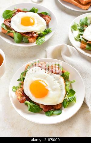 Offene Sandwiches mit Spiegeleiern, Speck und Gemüseblättern auf dem Teller auf hellem Steingrund. Stockfoto