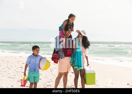 Glücklicher Birazialmann, der Tochter auf den Schultern trägt, während er mit Frau und Sohn am Strand spazierengeht Stockfoto